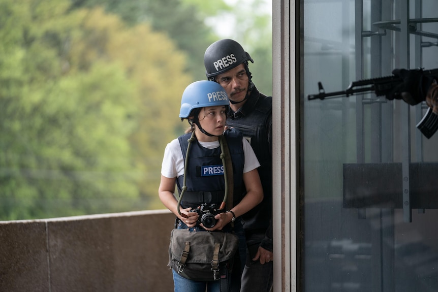 A man and young woman stare out from a balcony corner wearing press hard hats and vests. A gun tip enters the image from right.