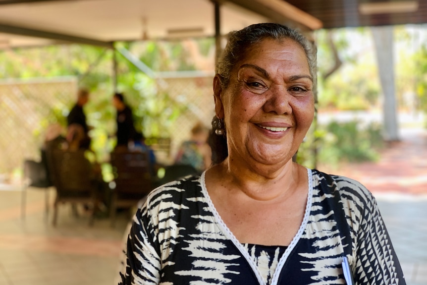 June Oscar in a patterned blouse smiles at the camera with tropical vegetation in the background.