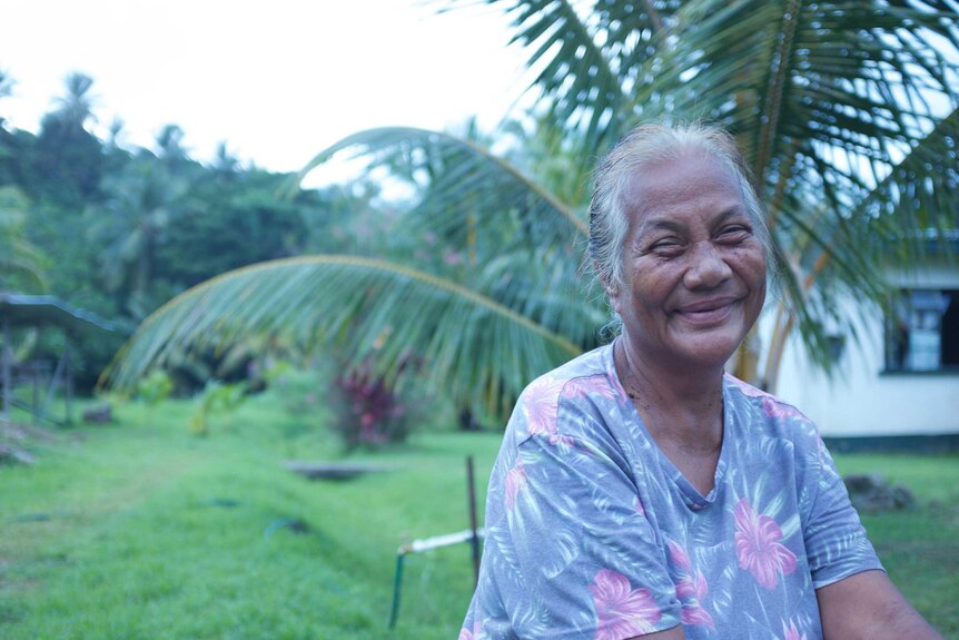 A woman smiles, sitting out the front of a house and palm-trees in Fiji.