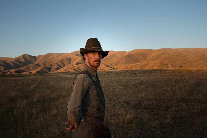 A 40-something man in a cowboy hat stands in a field, ringed by table hills, arms behind his back, looking into the camera lens