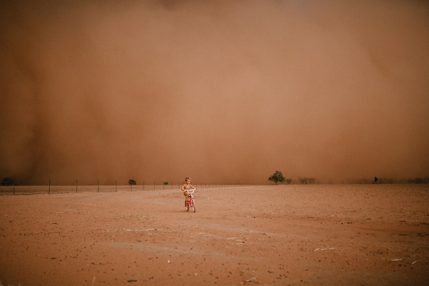 A small child rides a bike as a dust storm approaches.
