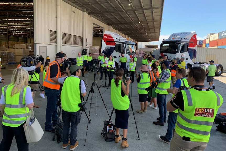A woman stands surrounded by reporters and camera crew in hi-vis vests.