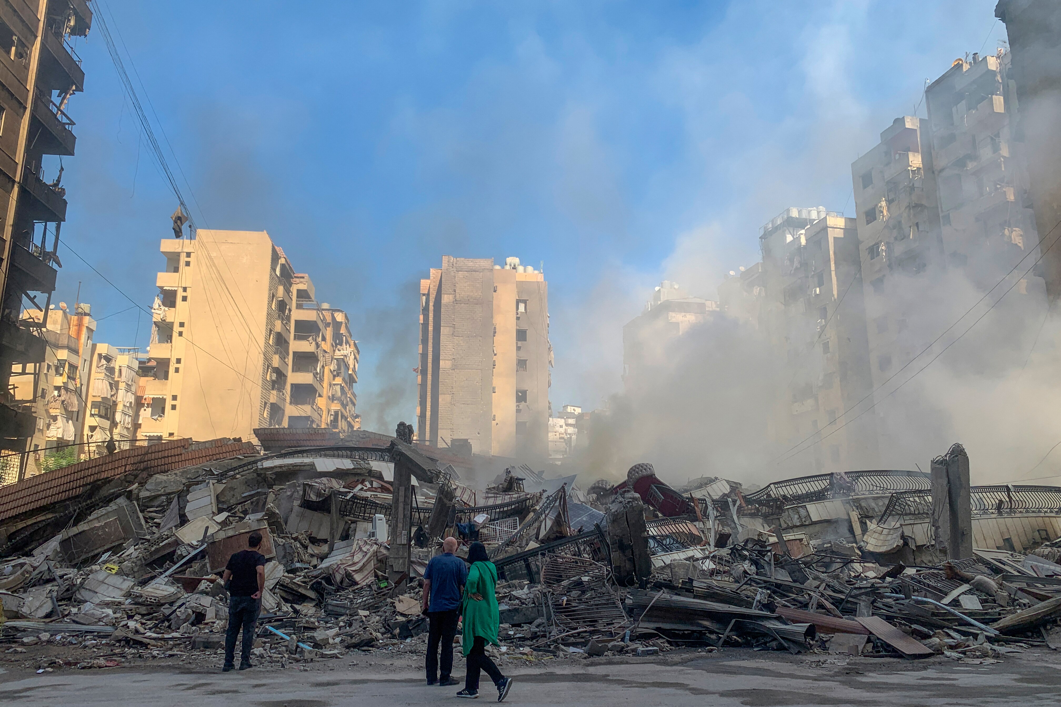 Three people stand in front of a smoking pile of debris, surrounded by buildings.