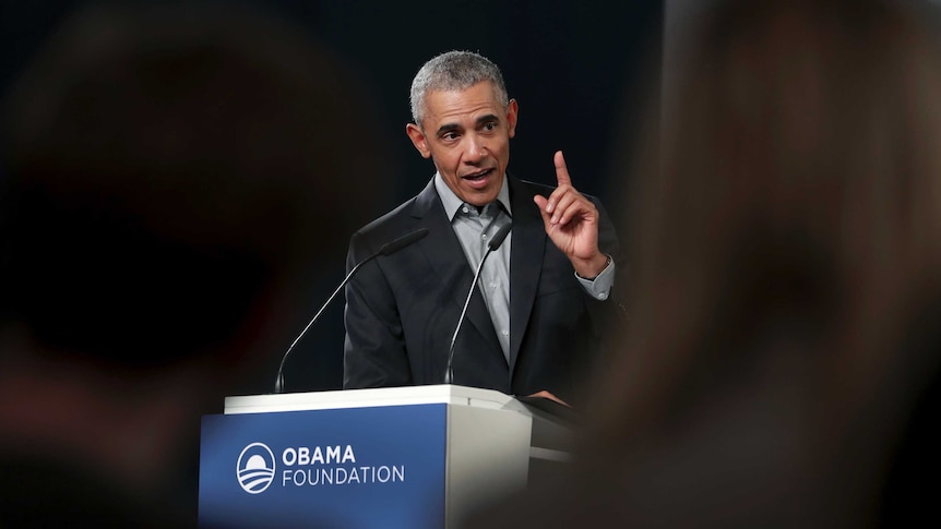 Former President Barack Obama holds up his index finger in a gesture as he speaks behind a podium.