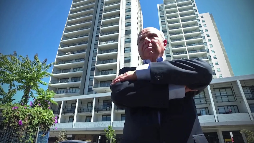 A man wearing a suit stands with his arms crossed. The photo is from a low angle, showing two large apartment blocks behind him.