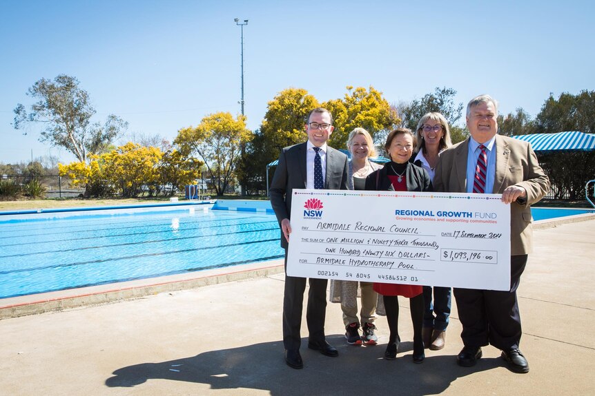 Three women and two men stand smiling, holding a large novelty cheque in front of a pool.