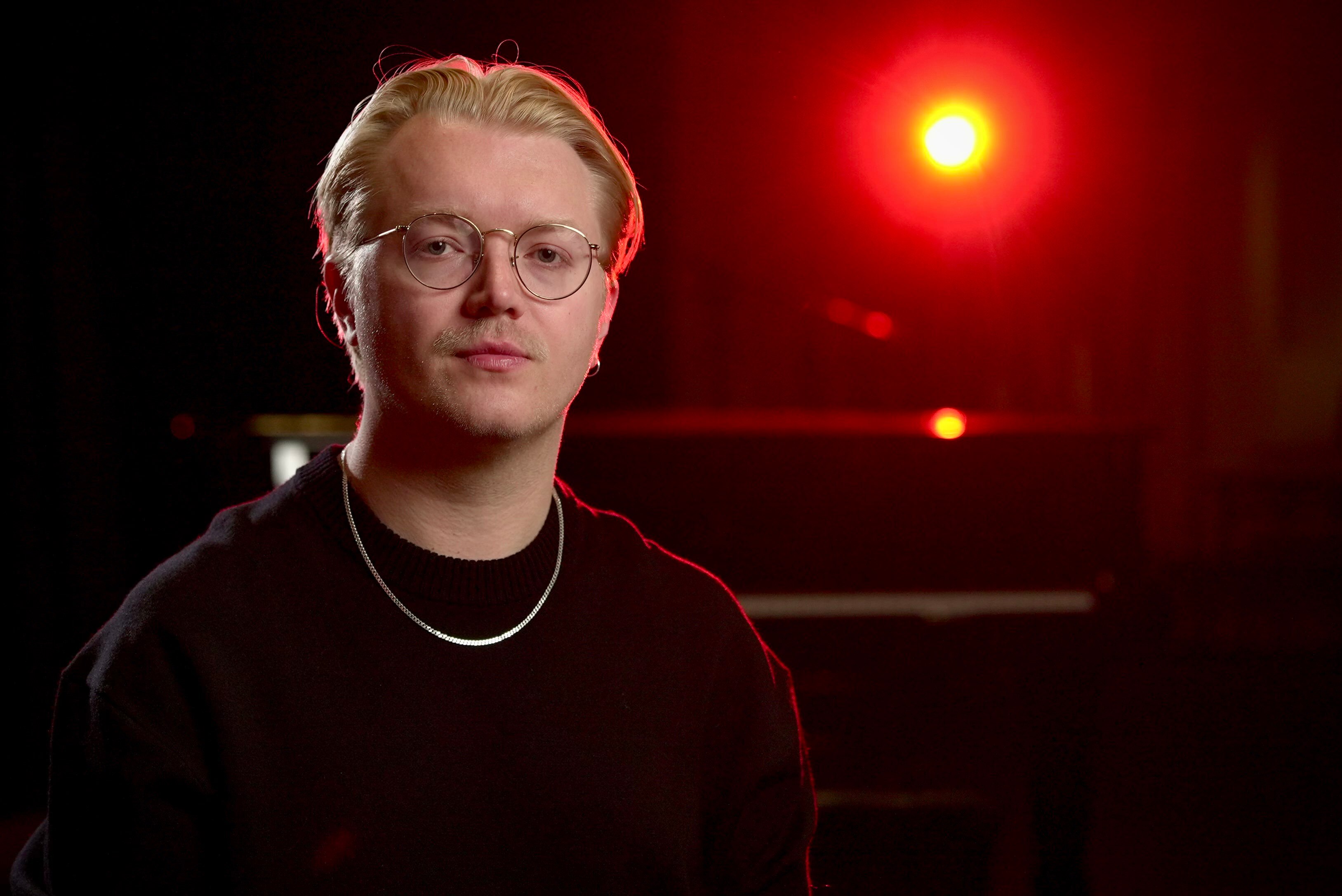 Un hombre con una camiseta oscura y gafas en un escenario a contraluz.