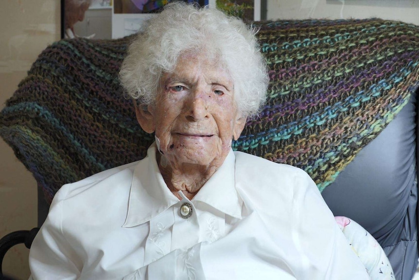 A head and shoulders image of an elderly woman in a white shirt sitting on an armchair with a crocheted throw over the back.