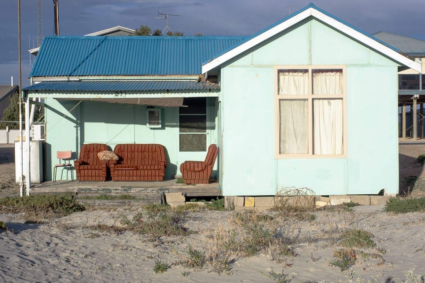 An old aquamarine-coloured beach shack with a blue corrugated iron roof and some comfortable-looking couches on the verandah.