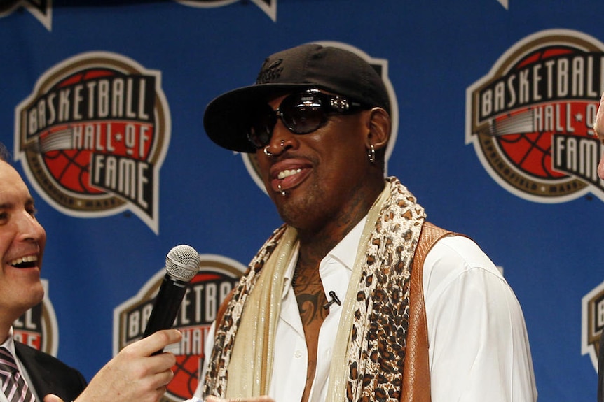 Rodman smiles. Someone holds a microphone up to his mouth.