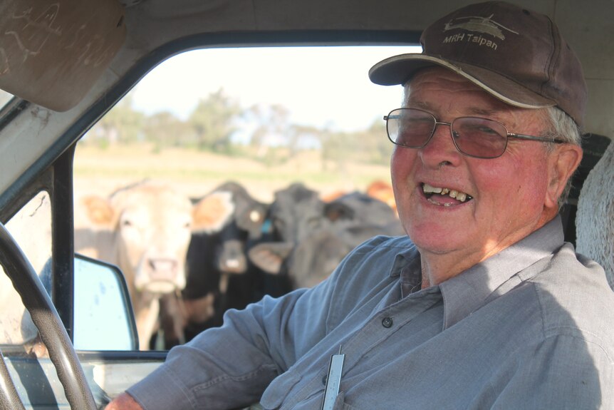A man is pictured wearing a hat and glasses. He smiles at the camera. Behind him are cows that curiously look on.