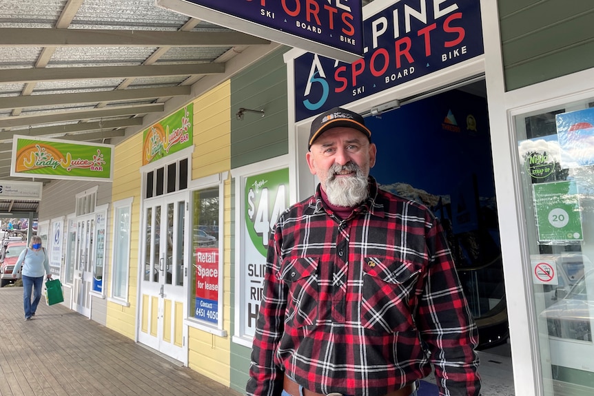 An older, bearded man standing out the front of a local alpine sports store wearing a dark cap and flannel shirt
