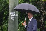 Albanese walks along a suburban street carrying an umbrella. Graffiti behind him reads 'have a bad ass day!'
