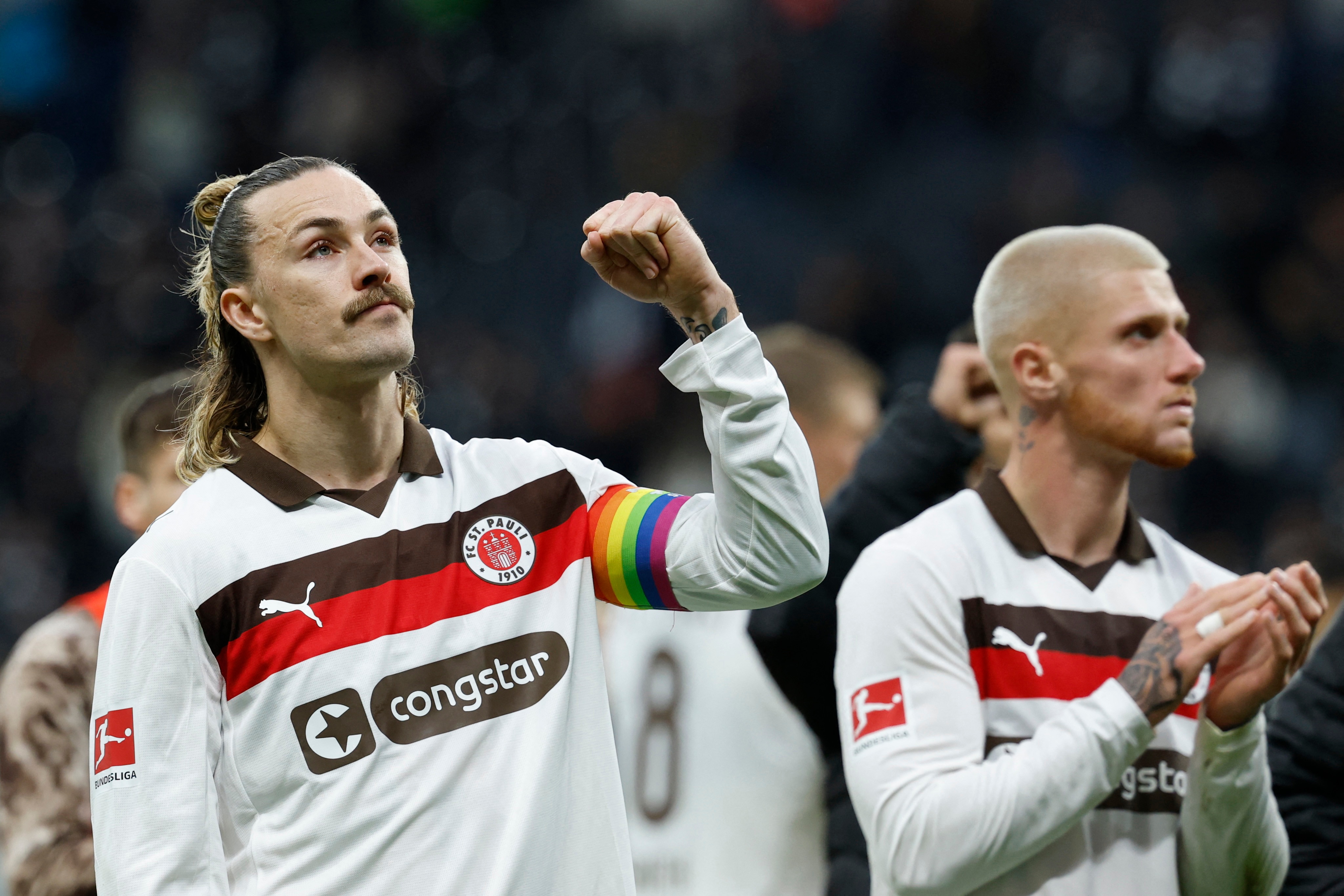 Surrounded by his St Pauli teammates, Irvine raises a fist. A Pride band is visible on his upper arm. 