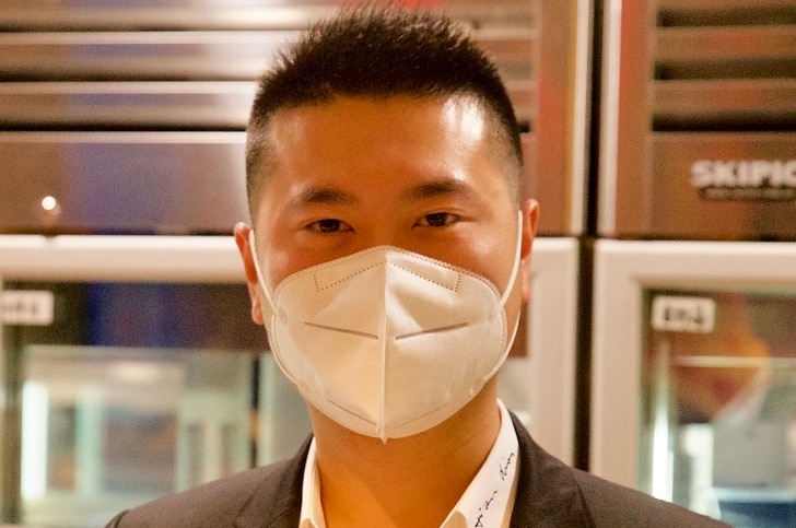 Yi Li stands behind the bench of his kitchen, wearing a face mask.