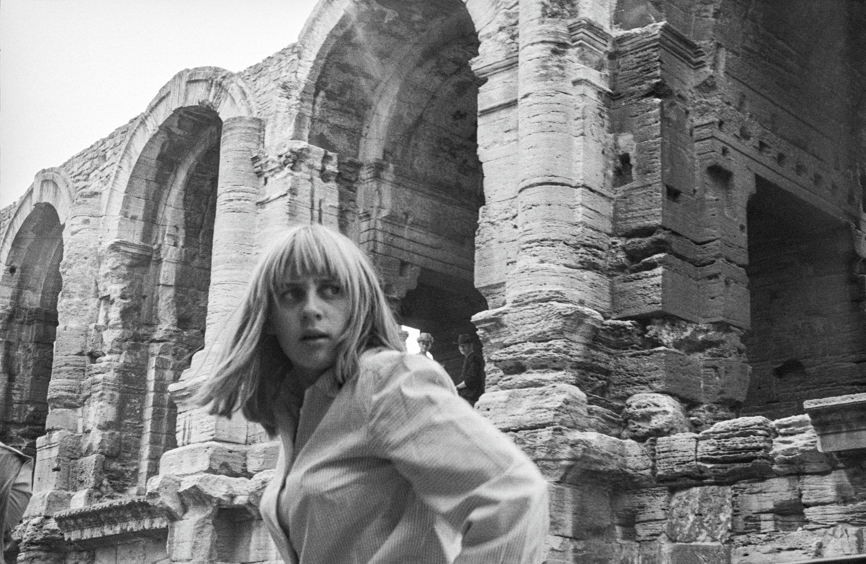 A B&W photo from the 60s of a blonde woman with a fringe, turning behind her, in front of a classical amphitheatre in France.