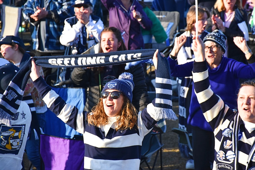 A woman holds up a Geelong Cats scarf as she cheers in a crowd.