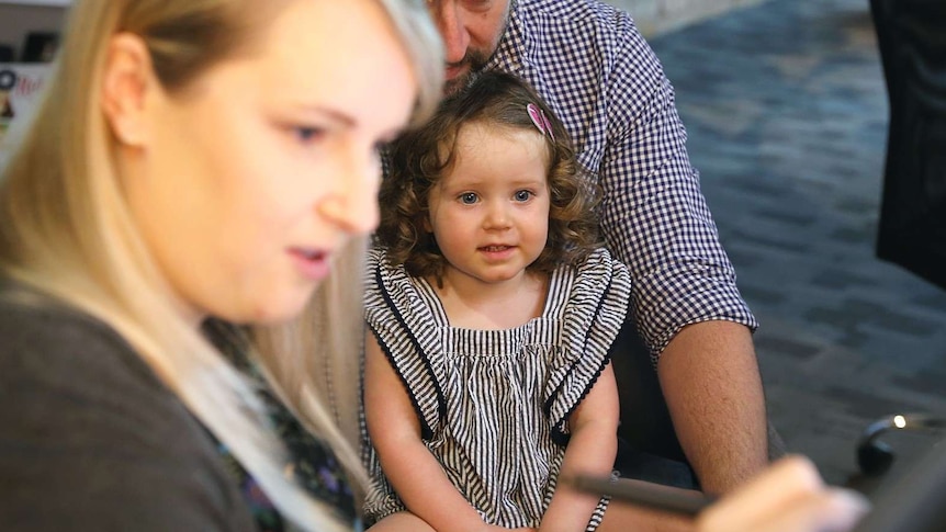 A girl sitting on her dad's lap and looking at a screen and smiling with an artist in the foreground.