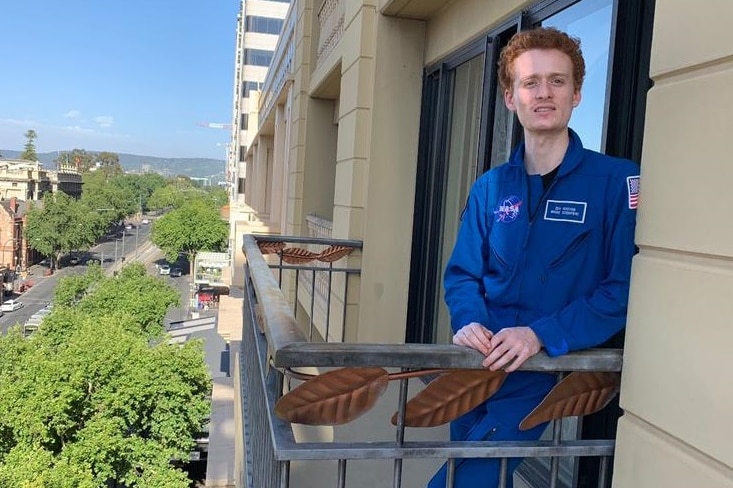A man wearing blue NASA coveralls standing on a hotel balcony
