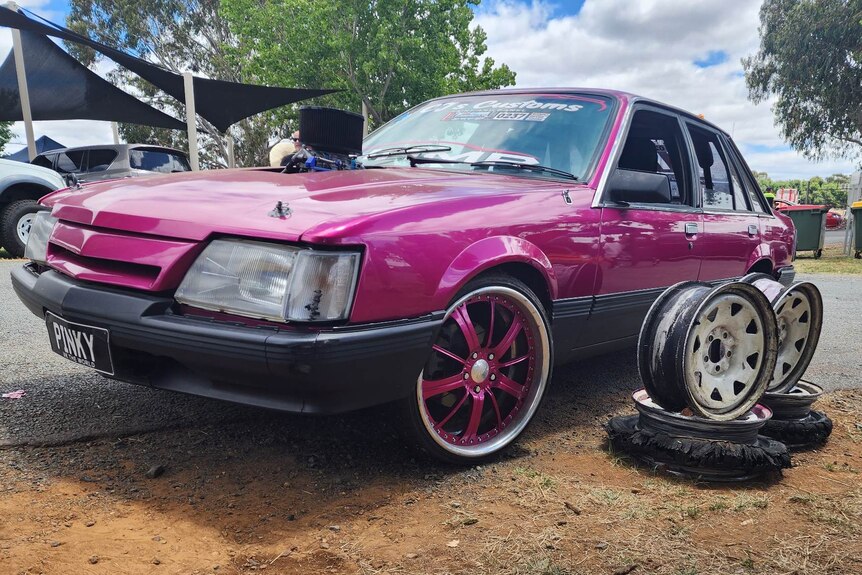 A bright pink 1986 Holden Commodore beside four shredded tyres following a burnout.