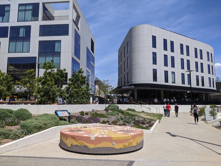 Tall modern curved buildings rise up as people walk past. An artwork is on display in the foreground.