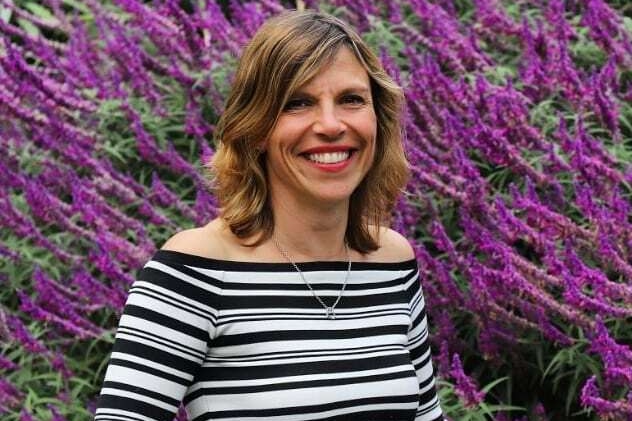 A woman wearing a black and white striped shirt stands in front of a garden with purple flowers