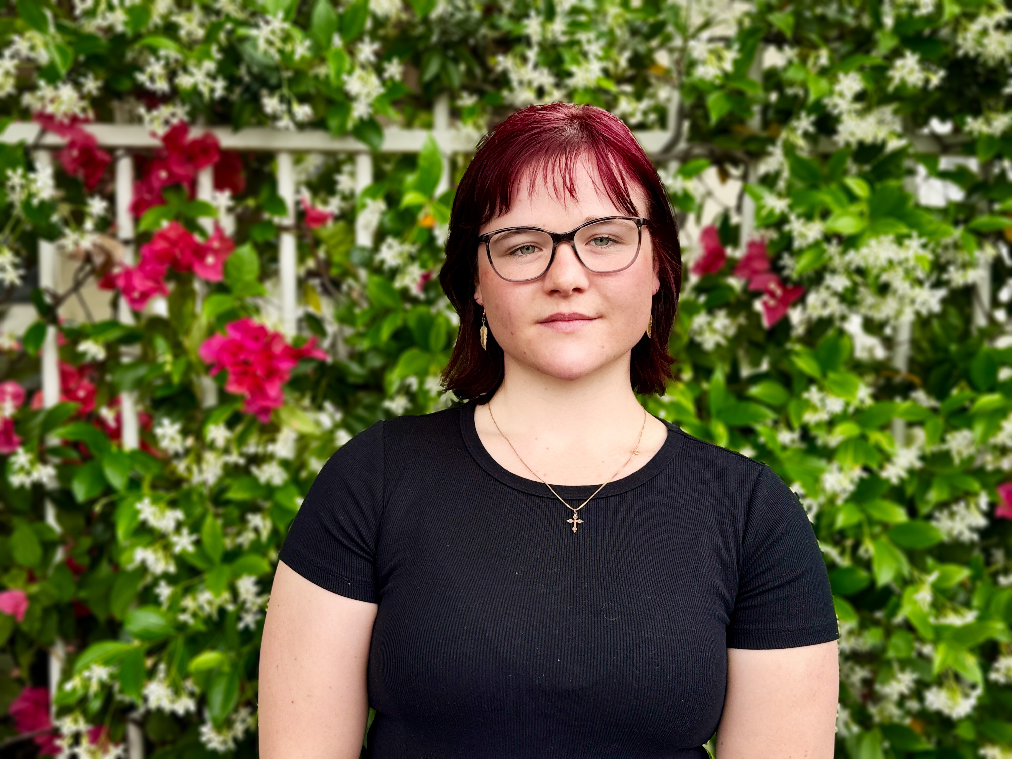 A young woman with short straight burgundy hair, black tshirt and glasses. Red flowers and shrub behind