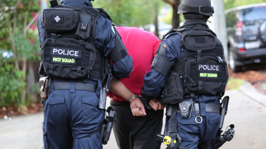 Man in red shirt and black tracksuit pants being led down a street in handcuffs by riot squad police officers.