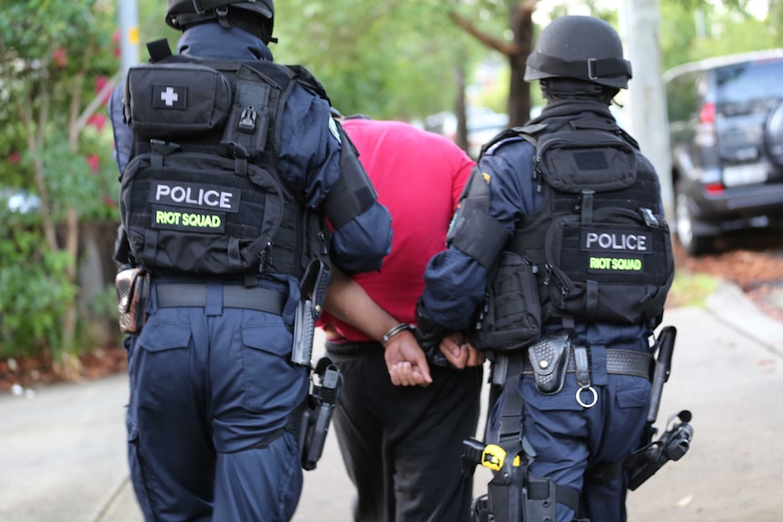 Man in red shirt and black tracksuit pants being led down a street in handcuffs by riot squad police officers.