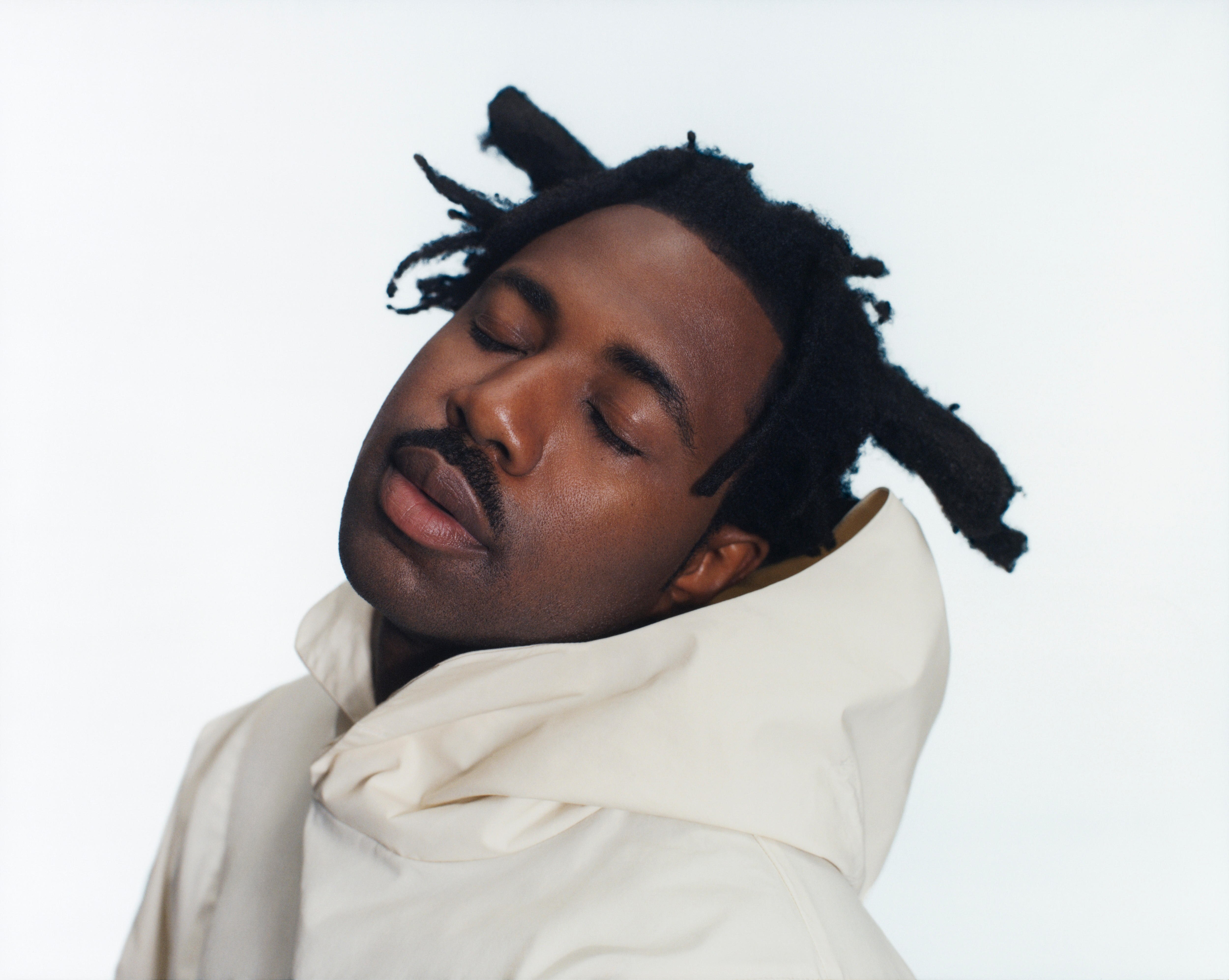 A press shot of Sampa tilting his head, eyes closed, wearing a white hoodie and sporting thick dreadlocks