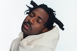 A press shot of Sampa tilting his head, eyes closed, wearing a white hoodie and sporting thick dreadlocks