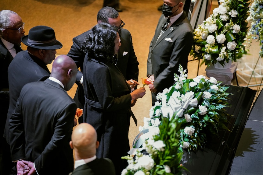 A woman holds a flower in front of a casket as other people stand nearby. 