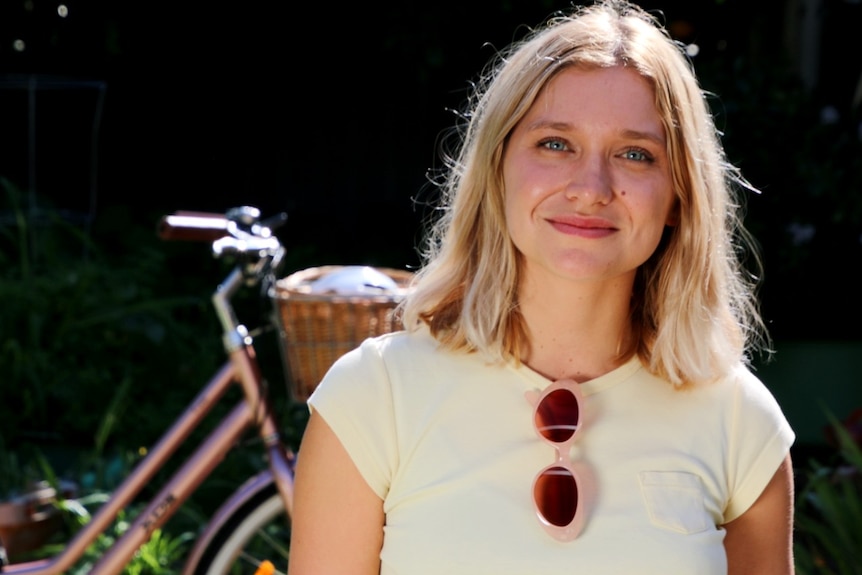 A girl with blonde hair and a pale yellow t-shirt with sunglasses folded over the neckline stands in front of a bicycle.