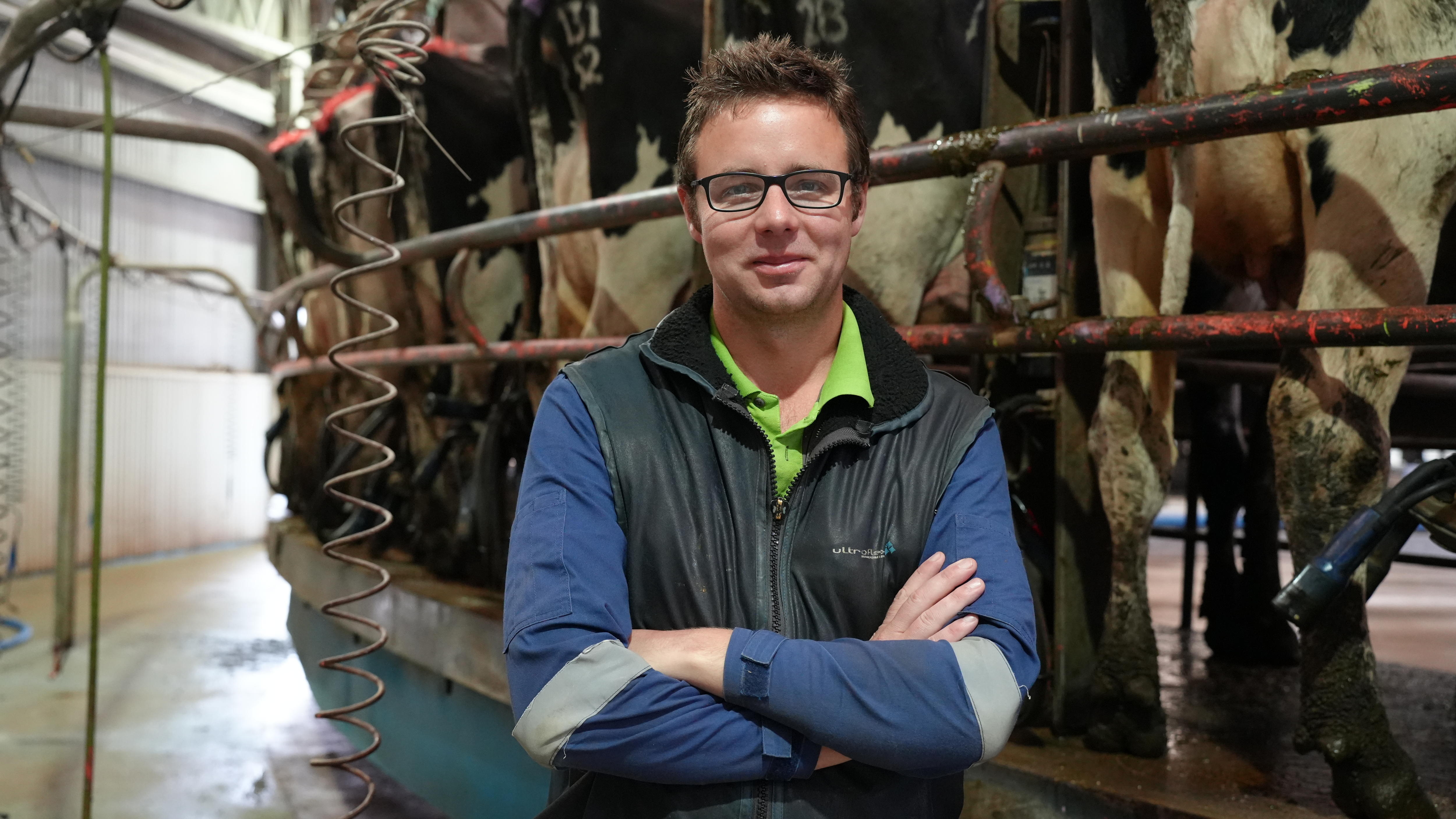 A dairy farmer stands with arms crossed with cows behind him in pens