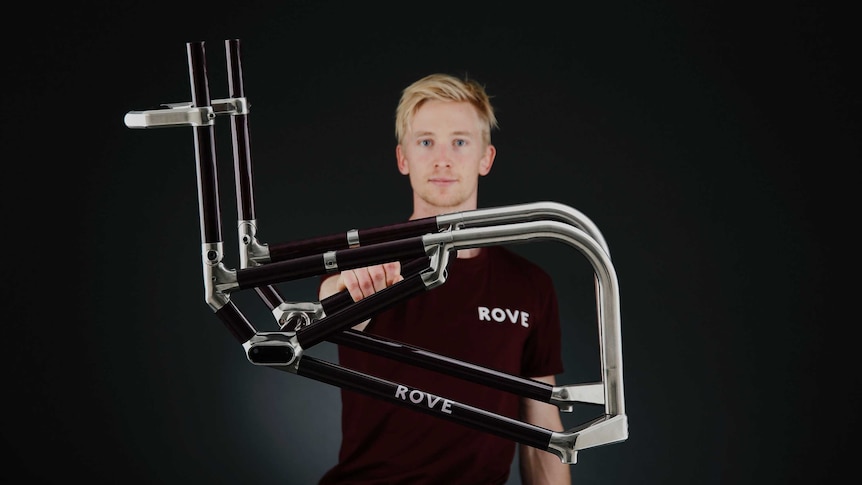 A young man holds up a wheelchair frame with one hand, demonstrating how light it is.