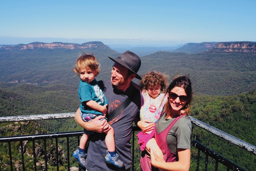 With a backdrop of lush bushland and mountains, a man and woman, both holding a small child, all squint in the sun, smiling.