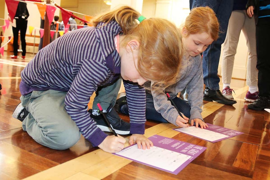 Two small girls kneel on the floor writing down the answers to the Great Easter Egg Trail questions on a piece of paper.