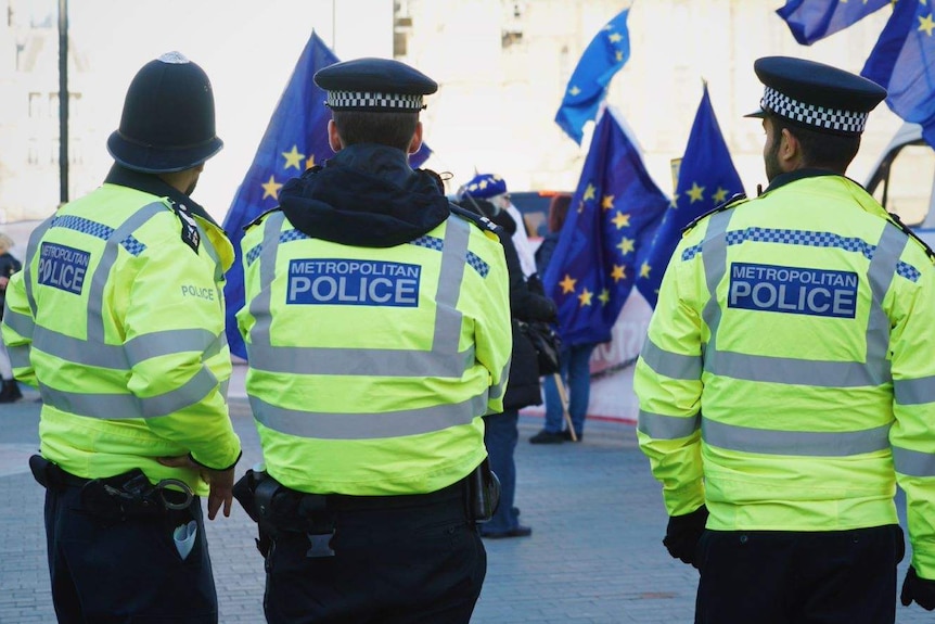 Three police officers wearing fluoro yellow jackets, with people waving European Union flags.