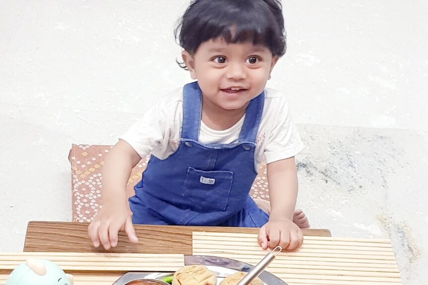 A boy wearing blue overalls and sitting in a high chair looks up fom his plate of food.