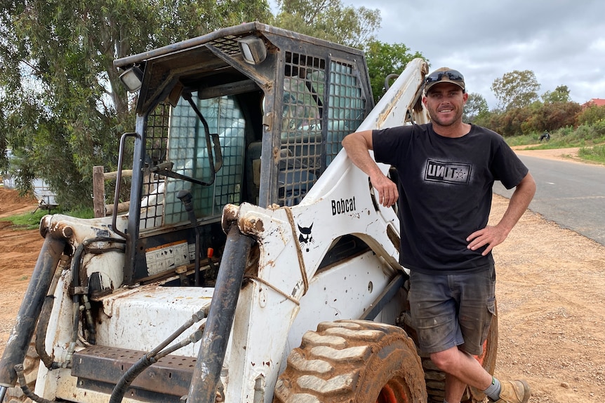 A man in a black shirt leans against a white bobcat digger