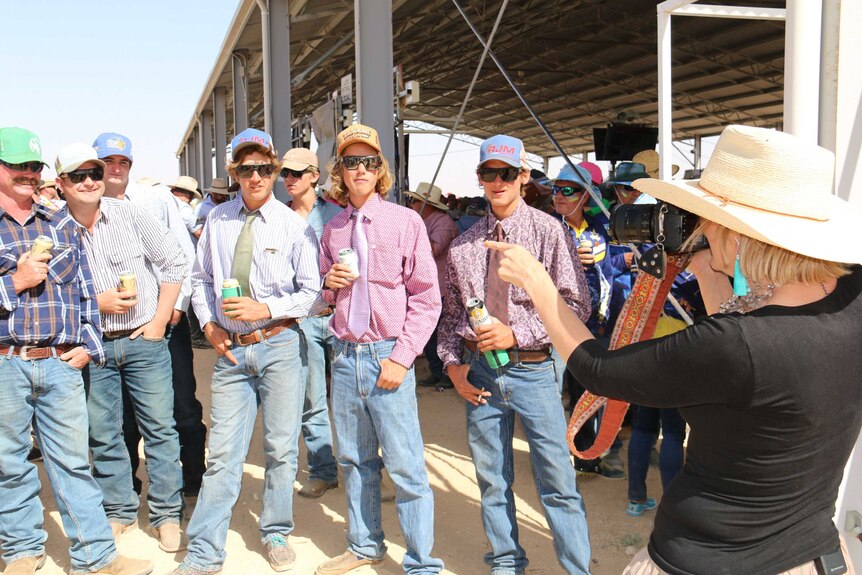 A woman wearing a wide brim hat takes a photo of a group of men all wearing jeans and dress shirt and hats
