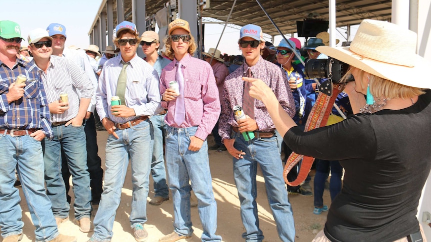 A woman wearing a wide brim hat takes a photo of a group of men all wearing jeans and dress shirt and hats