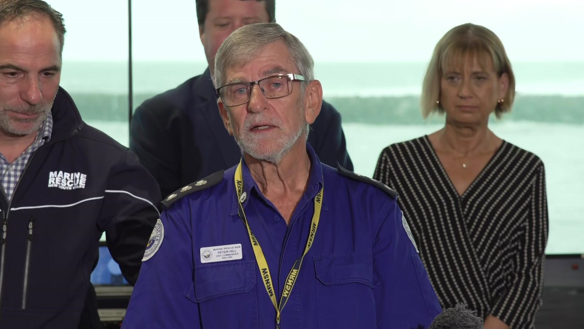 Man with grey hair and glasses in a blue Marine Rescue uniform with politicians behind him.