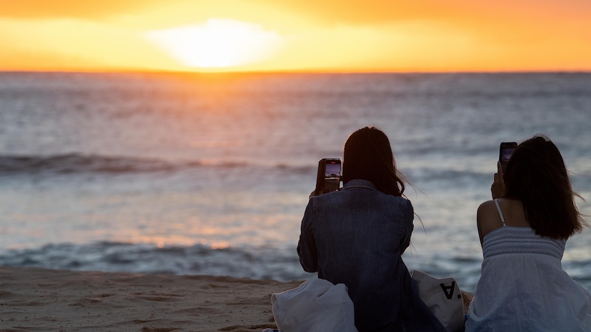 Two women sit on a beach taking photos on their phone of a sunrising over the water at Bondi Beach