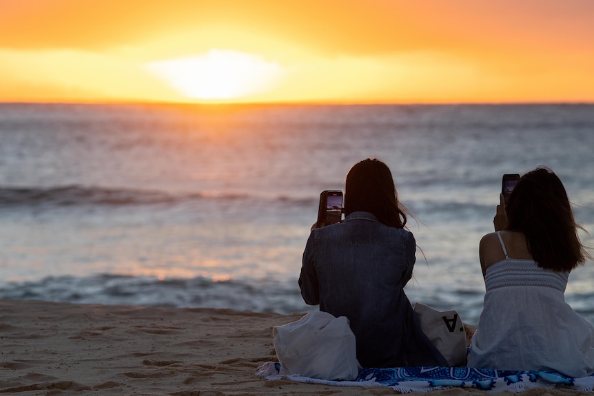 Two women sit on a beach taking photos on their phone of a sunrising over the water at Bondi Beach