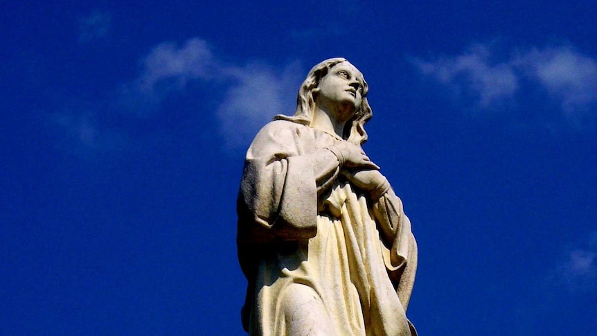 Against a rich blue sky, a statue of the Catholic Virgin Mary stands on an ornate plinth with children's heads beneath her feet.