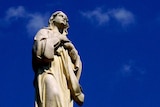Against a rich blue sky, a statue of the Catholic Virgin Mary stands on an ornate plinth with children's heads beneath her feet.