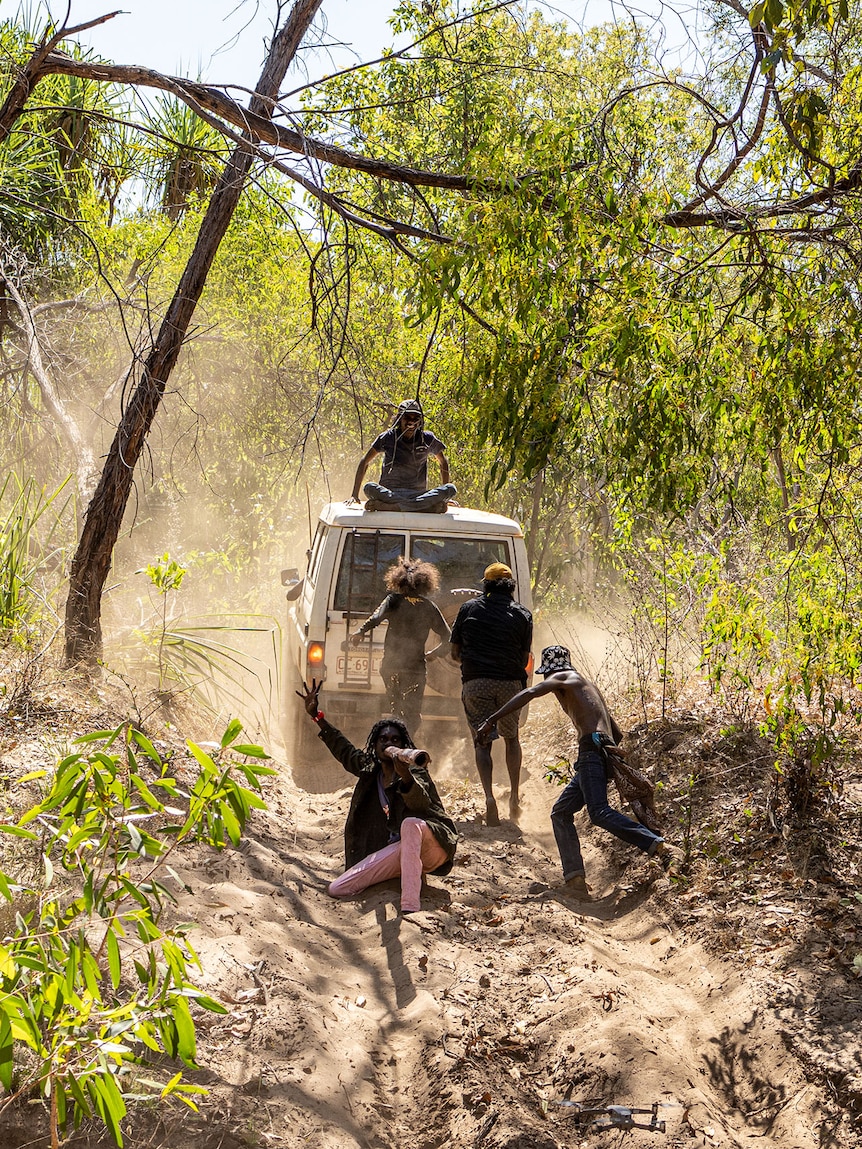 A white troupe drives over sand in the bush with five people sitting on the roof