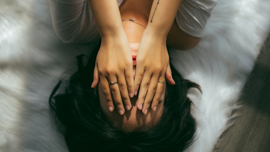 Bird's eye view of person with black hair in white shirt laying on white bed with hands covering face.