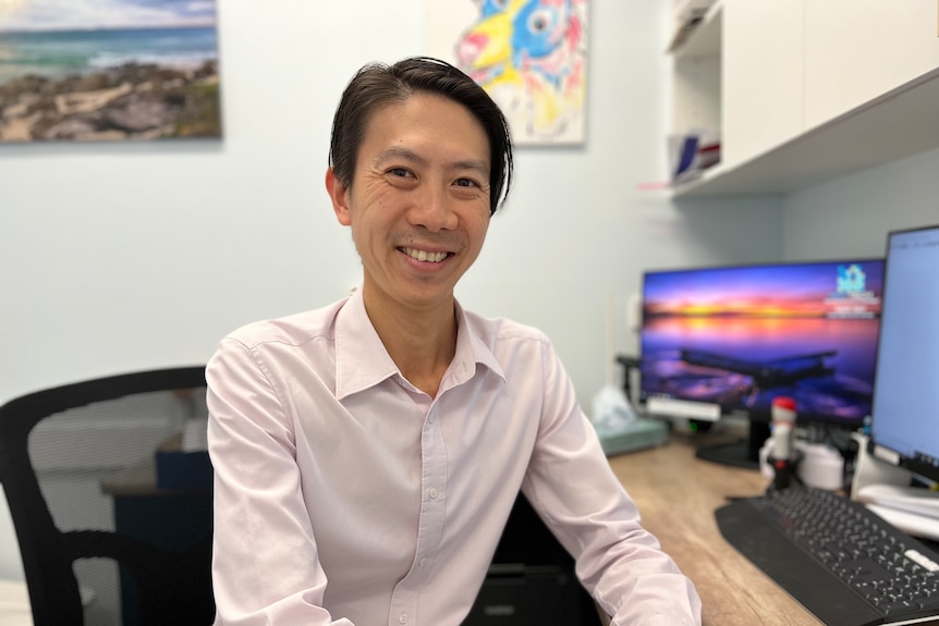 A doctor sits in his practice smiling at the camera.